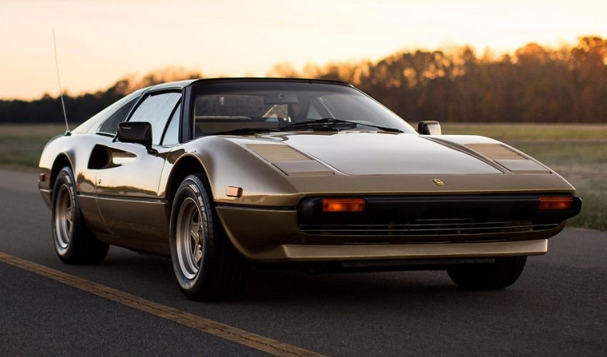 A classic brown Ferrari 308 GTS cruises down a rural road at sunset.