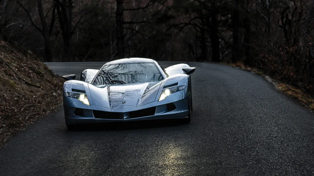 A sleek silver hypercar on a forest road at dusk.