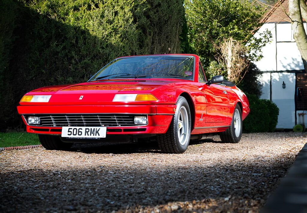 A red Ferrari 308 GTS parked on a gravel driveway with greenery and a building in the background.