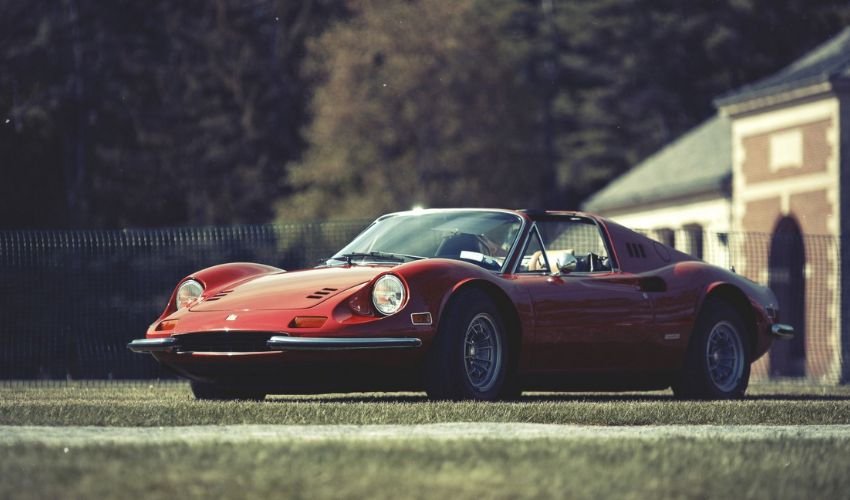 A classic red sports car parked on grass with a blurred building in the background.